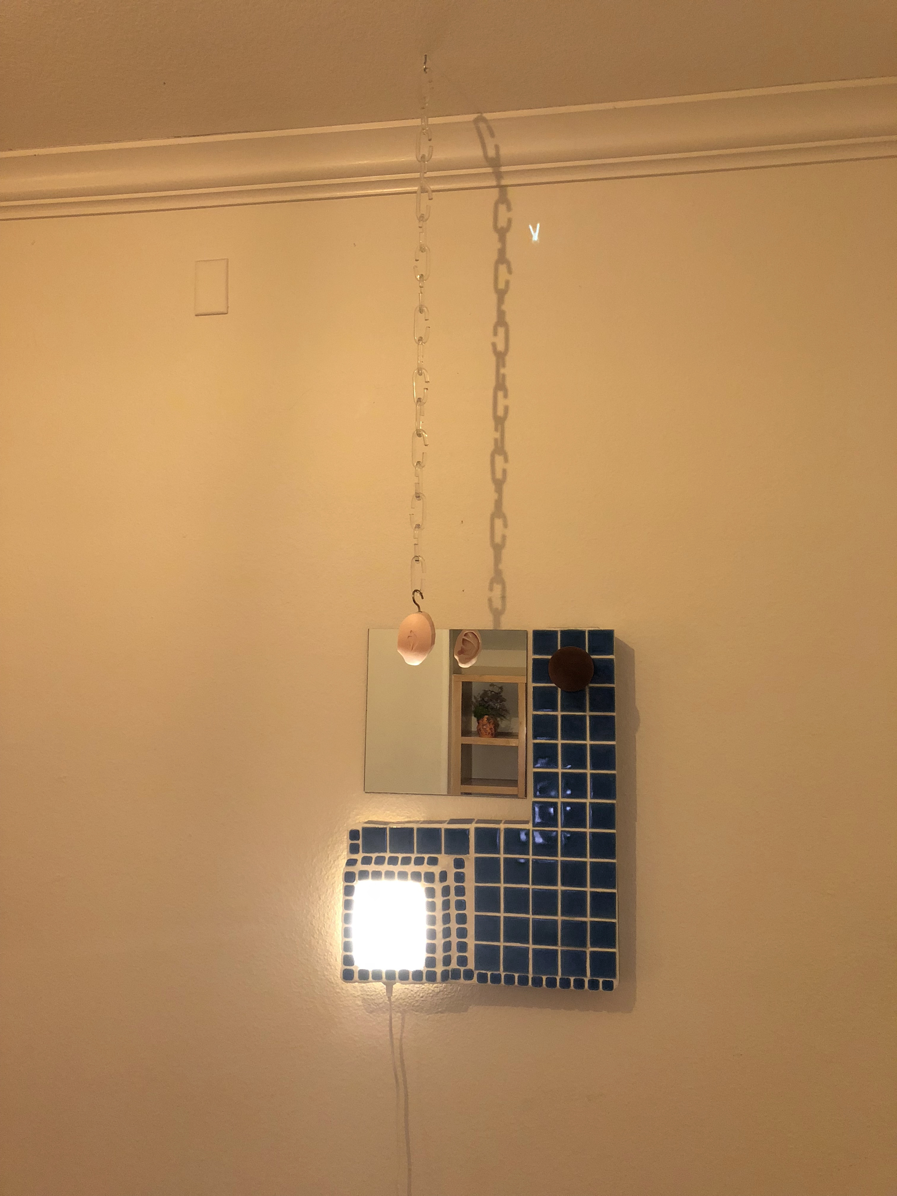 A sculptural wall lamp made of tile and mirror with a soap sculpture hung in front of it